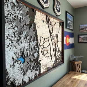 Könnte beinhalten: Eine gerahmte, mehrschichtige Holzkarte eines geografischen Gebiets mit detaillierten Konturlinien und einem blauen See. Die Karte ist in einem schwarzen Rahmen gefasst und hängt an einer grünen Wand, mit militärischen Emblemen und einer Colorado-Flagge im Hintergrund.