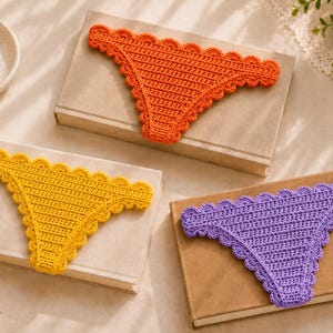 May include: Three triangular crochet coasters in orange, yellow, and purple, each featuring a scalloped edge border. The coasters are displayed individually on top of three separate rectangular books.