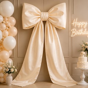 May include: A large cream-colored fabric bow centered on a wall, flanked by a vertical balloon garland in shades of cream and white. A neon sign reads Happy Birthday above a three-tiered cake on a pedestal, with floral arrangements nearby.