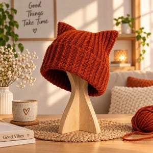 Può includere: Un berretto in maglia a coste color ruggine con due forme a punta di orecchie di gatto sulla parte superiore. Il cappello è esposto su un supporto in legno chiaro, che mette in risalto la trama verticale del filato e il bordo ripiegato.