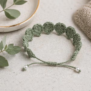 May include: A sage green crochet bracelet with a series of circular shapes. The bracelet has a drawstring closure with two small white pearl beads. The bracelet is on a neutral surface with a white and gold rimmed dish and a beige washcloth in the background.