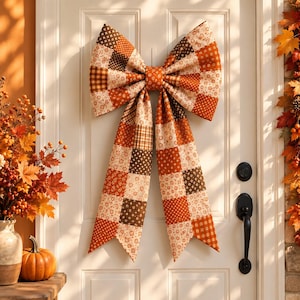 May include: A large decorative bow made of fabric with a patchwork design featuring various autumn-themed patterns in shades of orange, brown, cream, and white. The bow has two wide loops and two long, hanging tails, displayed on a white paneled door.