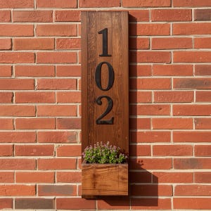 May include: A wooden address sign with the number "102" in black, mounted on a red brick wall. The sign has a built-in planter box filled with small purple flowers.
