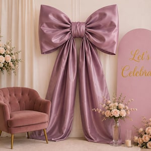 May include: A large mauve satin bow decoration hanging against a wall, accompanied by a mauve velvet armchair with gold legs and a pink arched sign featuring the gold text Let's Celebrate, surrounded by arrangements of light pink roses.