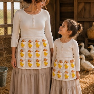 May include: Two cream-coloured crochet egg gathering aprons featuring a grid of square pockets, each adorned with a small yellow chick appliqué. The aprons are worn over long-sleeved cream shirts and long beige skirts, with brown eggs in pockets.