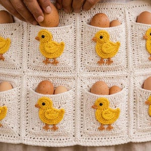 May include: A cream-coloured crochet apron featuring multiple square pockets, each adorned with a small, yellow crocheted chick appliqué. The pockets are designed to hold brown chicken eggs, with several eggs visible inside the individual compartments.