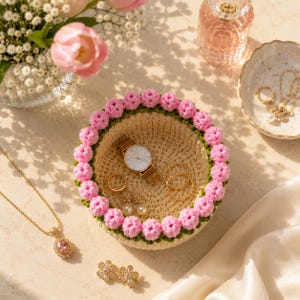 May include: A crocheted jewelry dish with a pink floral border holds a gold watch, rings, and earrings. A gold necklace and flower earrings are also displayed. A vase of pink tulips and a perfume bottle are in the background.