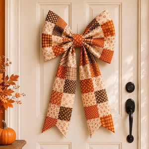 Könnte beinhalten: Eine große dekorative Schleife aus Stoff mit einem Patchwork-Design, das verschiedene Muster in Orange-, Braun- und Cremetönen aufweist. Die Schleife besteht aus zwei breiten Schlaufen und zwei langen, herabhängenden Bändern aus Stoffquadraten.