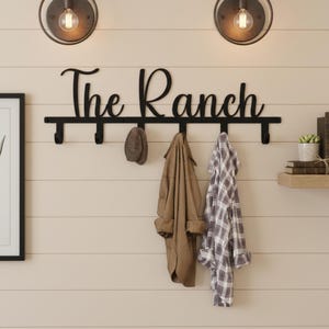 May include: A black metal wall-mounted coat rack with the cursive text "The Ranch". The rack has several hooks and is adorned with a hat and two garments. Two vintage-style light fixtures are above the rack.