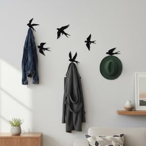 Op de afbeelding: Zwarte metalen vogelvormige wandhaken met een spijkerjasje, een grijze jas en een groene hoed eraan. De haken zijn in een decoratief patroon gerangschikt, met extra vogel silhouetten die op de muur vliegen.