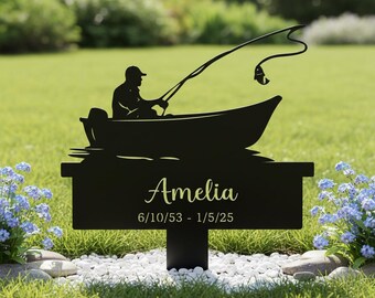 May include: Black metal memorial sign featuring a silhouette of a person fishing from a boat. The sign includes the name "Amelia" and dates 6/10/53 - 1/5/25. The sign is set in a garden with flowers and white stones.