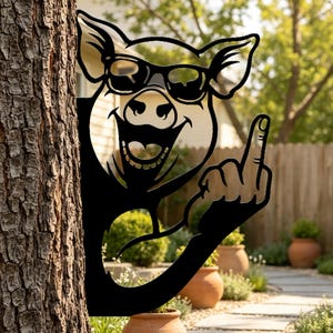 May include: A black metal garden silhouette of a pig wearing sunglasses and smiling, with one hand raised showing the middle finger. The cutout design is positioned against the side of a tree trunk, creating a decorative outdoor accent piece.