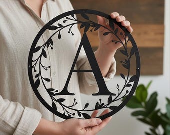 May include: Black metal wall art featuring the letter "A" within a circular frame. The frame is decorated with a leaf and berry design. The decorative piece is held up by a person.
