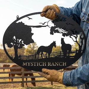 Pode incluir: Placa oval de metal preto com uma cena de rancho com dois cavalos, árvores e uma cerca. A placa tem as palavras "MYSTICH RANCH" em letras douradas. A placa é segurada por uma pessoa usando luvas.