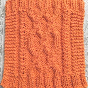 May include: An orange knitted square with a cable knit design. The square features a central cable pattern flanked by textured columns and ribbed edges. The yarn is a vibrant orange color, and the stitches are clearly defined.