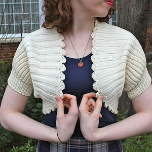 May include: A white crocheted shrug with a scalloped edge. The shrug is worn over a dark blue top and a heart-shaped pendant necklace.