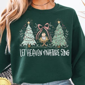 May include: Dark green sweatshirt with a Christmas-themed graphic. The design features two Christmas trees, a pink bow, and a baby in a manger. The text "Let Heaven & Nature Sing" is printed in white.