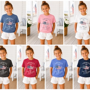 May include: A collection of t-shirts in various colors, each featuring the word "AMERICA", a stylized American flag, and the text "250 YEARS". The shirts are worn by a young person and paired with white shorts.