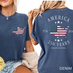 May include: Denim blue t-shirt with a distressed American flag design in the shape of the United States. The back of the shirt features the word "AMERICA", the years "1776" and "2026", and "250 YEARS SEMIQUINCENTENNIAL".