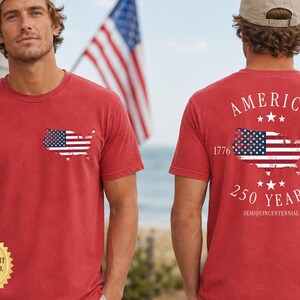 May include: Red t-shirts with an American flag design. One shirt has a small flag map on the chest. The other shirt has a larger flag map with the words "AMERICA", "1776", "2026", and "250 YEARS".