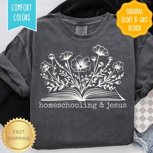 May include: Dark gray long-sleeve shirt with a white floral design emerging from an open book. The text "homeschooling & jesus" is below the book. The shirt has "Comfort Colors" and "Original Glory & Grit Design" labels.