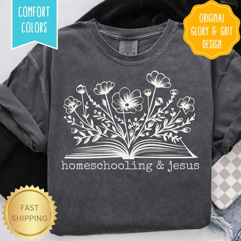 May include: Dark gray long-sleeve shirt with a white floral design emerging from an open book. The text "homeschooling & jesus" is below the book. The shirt has "Comfort Colors" and "Original Glory & Grit Design" labels.