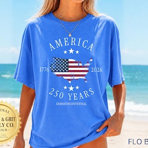 May include: A blue t-shirt with the word "AMERICA" arched across the top and the years "1776" and "2026" flanking a map of the United States with a flag design. The text "250 YEARS" is below the map, with "SEMIQUINCENTENNIAL" below that. The shirt is labeled "FLO BLUE".