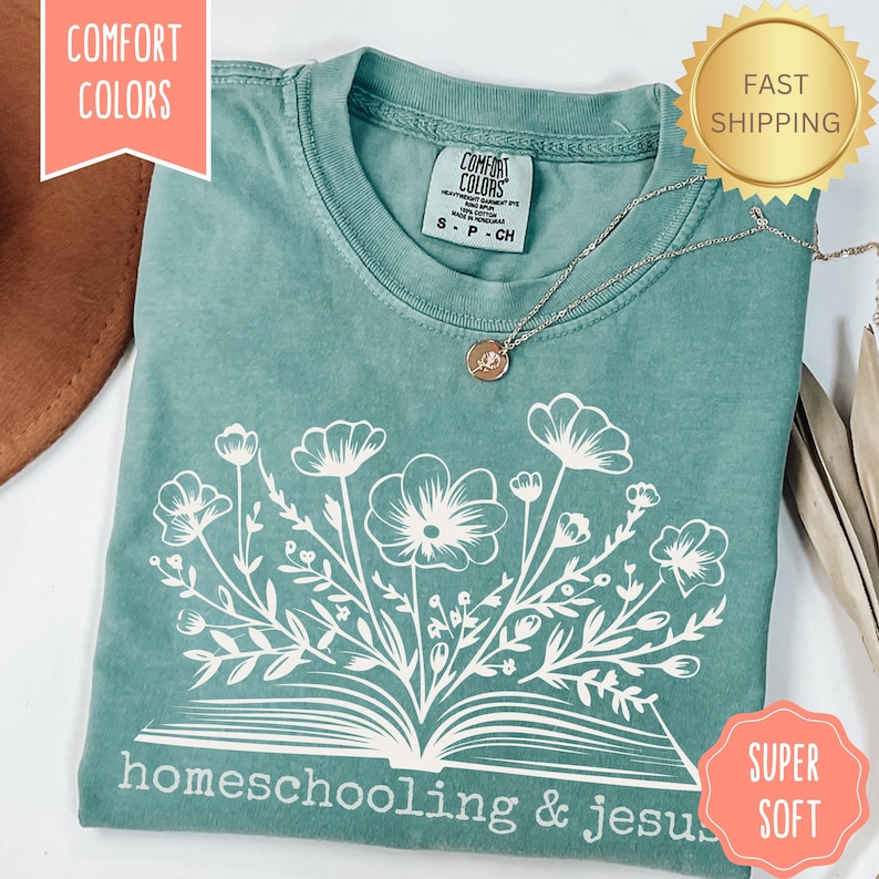 May include: A teal Comfort Colors t-shirt with a white floral design over an open book, and the words "homeschooling & jesus". A gold pendant necklace rests on the shirt. The shirt is labeled "Super Soft" and "Fast Shipping".