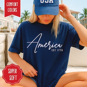 May include: Navy blue t-shirt with the word "America" in white script and "EST 1776" below it. The model is wearing a blue baseball cap with "USA" on it. The shirt has a "Super Soft" sticker.