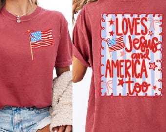 May include: Crimson-colored t-shirt featuring an American flag graphic on the front and the text "LOVES Jesus and AMERICA too" on the back. The design includes stars, stripes, and decorative elements.