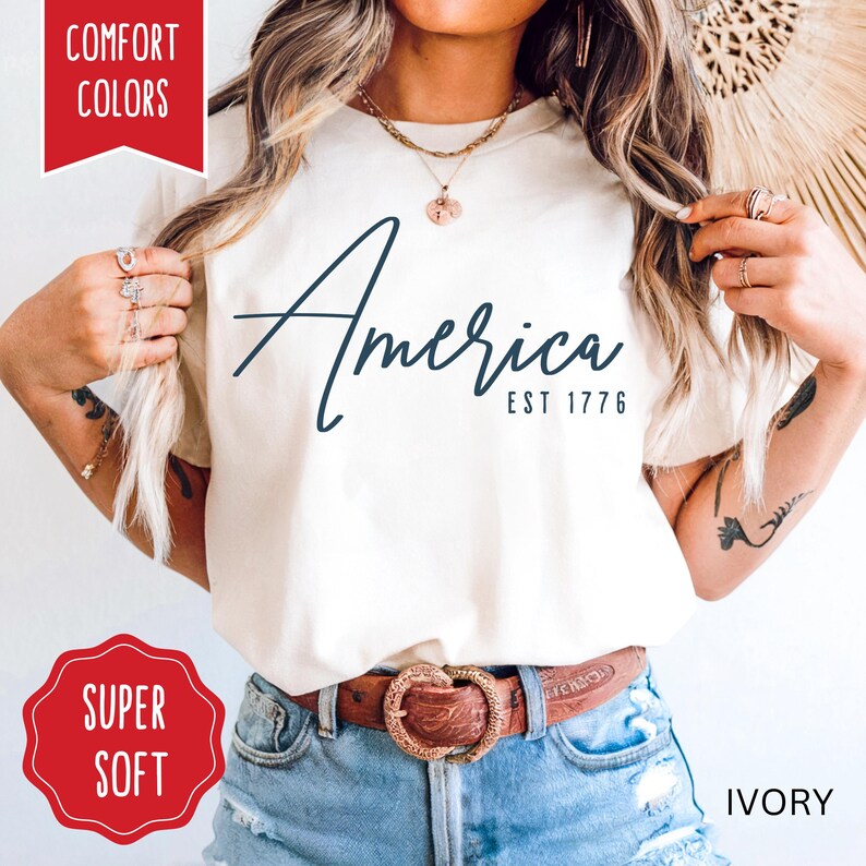 May include: Ivory-colored t-shirt with the word "America" in navy script and "EST 1776" below. The shirt has a crew neck and short sleeves. The words "Comfort Colors" and "Super Soft" are in red boxes.