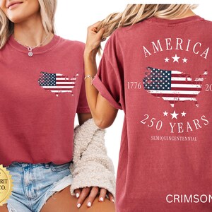 May include: Crimson-colored t-shirt featuring an American flag design. The front has a small flag outline, while the back displays "AMERICA" above a flag map, "1776-2026", and "250 YEARS". The shirt is a comfortable, casual style.