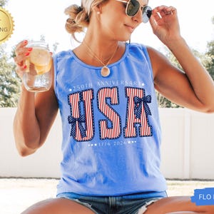 May include: A light blue tank top with the word "USA" in red and white stripes, with blue bows. The text "250th Anniversary 1776-2026" is also printed on the shirt. A gold "Fast Shipping" sticker is in the upper left corner.