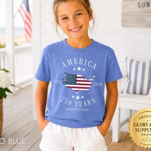 May include: A Flo Blue t-shirt with the word "AMERICA" and the years "1776" and "2026" printed on it. The shirt features a map of the United States with the American flag design. The text "250 YEARS SEMIQUINCENTENNIAL" is also printed on the shirt.