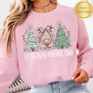 May include: Pink sweatshirt with a Christmas-themed graphic. The design features two decorated Christmas trees, a baby in a manger, and the text "LET HEAVEN & NATURE SING". The sweatshirt has a crew neck and long sleeves.