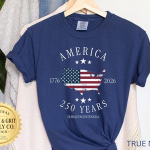 May include: A navy blue t-shirt with the word "AMERICA" above a map of the United States, featuring the American flag. The shirt also displays the years "1776" and "2026", and the text "250 YEARS SEMIQUINCENTENNIAL".