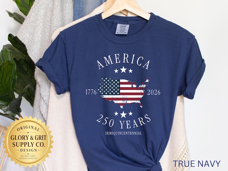 May include: A navy blue t-shirt with the word "AMERICA" above a map of the United States, featuring the American flag. The shirt also displays the years "1776" and "2026", and the text "250 YEARS SEMIQUINCENTENNIAL".