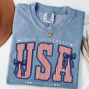 May include: A light blue t-shirt with the word "USA" in red and white stripes, accented with blue bows and stars. The shirt commemorates the 250th anniversary, with the years 1776-2026 printed below. A gold necklace rests on the shirt.