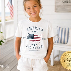 May include: A white t-shirt with the word "AMERICA" above a graphic of the American flag within a map of the United States. The text "1776" and "2026" flank the flag, with "250 YEARS" below. The shirt is worn by a young person.