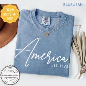 May include: A blue jean colored t-shirt with the word "America" in white script, and "EST 1776" below it. The shirt has a crew neck and a small tag. A gold necklace with a pendant and a dried plant are also visible.