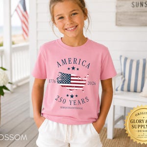May include: A pink t-shirt with the word "AMERICA" above a map of the United States with the American flag inside. The shirt also has the text "1776 2026 250 YEARS SEMIQUINCENTENNIAL". The shirt is worn by a young person.