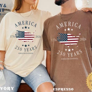 May include: Two t-shirts, one ivory and one espresso, each with a graphic celebrating America's 250th anniversary. The design includes the word "AMERICA", a map of the USA with the American flag, and the years 1776 and 2026.