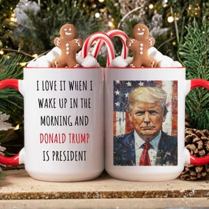 K&ouml;nnte beinhalten: Zwei wei&szlig;e Keramikbecher mit roten Griffen, mit dem Text "I LOVE IT WHEN I WAKE UP IN THE MORNING AND DONALD TRUMP IS PRESIDENT." Ein Becher hat ein Portr&auml;t von Donald Trump. Lebkuchenm&auml;nner und Zuckerstangen schm&uuml;cken die Becher.
