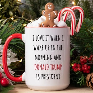 K&ouml;nnte beinhalten: Wei&szlig;e Keramiktasse mit rotem Henkel und Rand, gef&uuml;llt mit Marshmallows. Ein Lebkuchenmann und zwei Zuckerstangen liegen obenauf. Die Tasse tr&auml;gt den Text "I LOVE IT WHEN I WAKE UP IN THE MORNING AND DONALD TRUMP IS PRESIDENT."