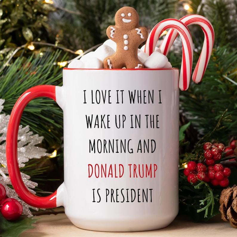 K&ouml;nnte beinhalten: Wei&szlig;e Keramiktasse mit rotem Henkel und Rand, gef&uuml;llt mit Marshmallows. Ein Lebkuchenmann und zwei Zuckerstangen liegen obenauf. Die Tasse tr&auml;gt den Text "I LOVE IT WHEN I WAKE UP IN THE MORNING AND DONALD TRUMP IS PRESIDENT."