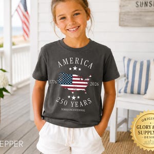May include: A dark gray t-shirt with the word "AMERICA" above a map of the United States with the American flag inside. The shirt also features the text "1776 2026 250 YEARS SEMIQUINCENTENNIAL". The shirt is worn by a young person.