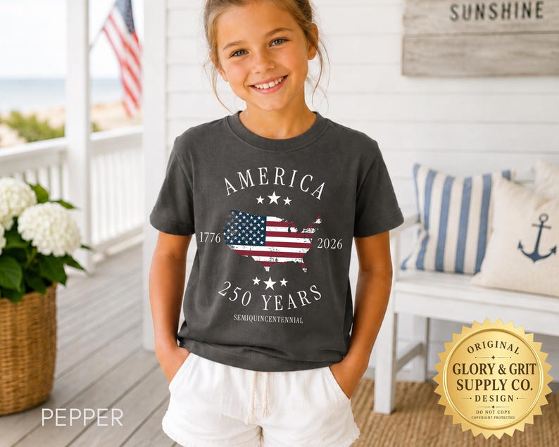 May include: A dark gray t-shirt with the word "AMERICA" above a map of the United States with the American flag inside. The shirt also features the text "1776 2026 250 YEARS SEMIQUINCENTENNIAL". The shirt is worn by a young person.