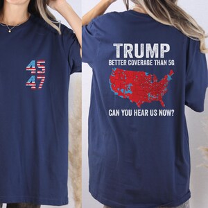 May include: A navy blue t-shirt with a red, white, and blue American flag design. The shirt has the numbers "45" and "47" on the left side and the text "TRUMP BETTER COVERAGE THAN 5G CAN YOU HEAR US NOW?" on the right side.