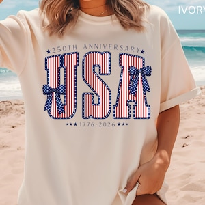 May include: Ivory-colored t-shirt with a patriotic design. The design features the letters "USA" in red and white stripes, outlined in blue, with blue bow accents. The text "250th Anniversary 1776-2026" is also included.