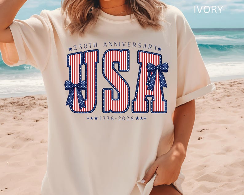 May include: Ivory-colored t-shirt with a patriotic design. The design features the letters "USA" in red and white stripes, outlined in blue, with blue bow accents. The text "250th Anniversary 1776-2026" is also included.